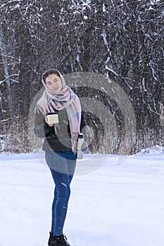 Young woman in warm shawl in winter snowy forest drinks hot tea. Full-length portrait,  Mug and thermos in his hands, hot drink