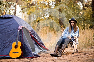 Young woman in warm sweater and black hat resting with guitar near camping tent in wilderness forest