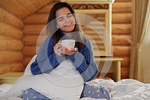 Young woman in warm sweater with cup of drink on bed at home
