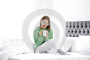 Young woman in warm sweater with hot drink on bed