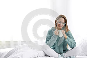 Young woman in warm sweater with hot drink on bed. Space for text
