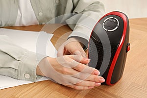 Young woman warming hands near compact electric heater at wooden table, closeup