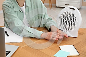Young woman warming hands near modern electric fan heater at wooden table indoors, closeup