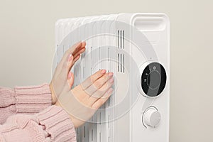 Young woman warming hands near modern electric heater on beige background, closeup