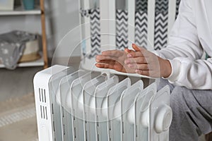 Young woman warming hands near modern electric heater indoors, closeup