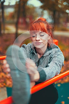 Young woman warming up and stretching her leg on bar in park at autumn, eyes closed