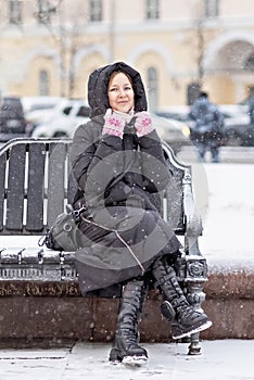 A young woman warmly dressed is sitting on a bench in the city on the street on a snowy cold day