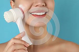 Young woman washing face with brush and foam on light blue background, closeup. Cosmetic products