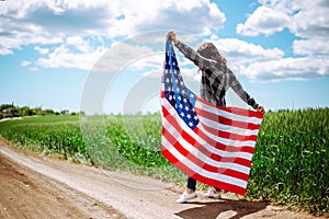 Young woman waves an american flag on the green wheat field. Patriotic holiday celebration. United States of America