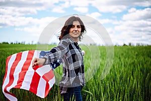 Young woman waves an american flag on the green wheat field. Patriotic holiday celebration. United States of America