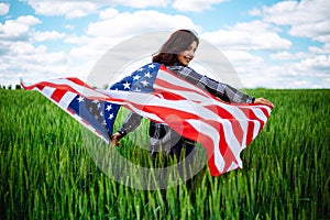 Young woman waves an american flag on the green wheat field. Patriotic holiday celebration. United States of America