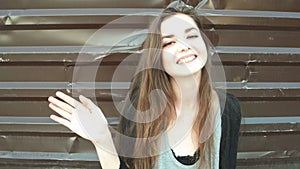 Young woman waving hand saying hello against metal fence of brown color. Smiling happy cheerful caucasian girl having