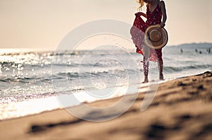Young woman in waving red summer dress walking on beach holding fashionable straw hat. Vacation, holiday, lifestyle concept