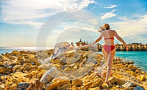 Young woman waving scarf in wind at beach. Happy woman in red bikini holding tissue and looking away at sea.