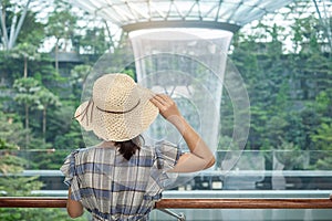 Young woman wear blue dress and hat, Asian traveler standing and looking to beautiful rain vortex at Jewel Changi Airport,