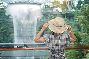 Young woman wear blue dress and hat, Asian traveler standing and looking to beautiful rain vortex at Jewel Changi Airport,