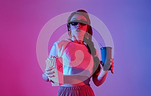 Young woman wearing 3D glasses holding popcorn and beverage watching exciting movie isolated on neon background