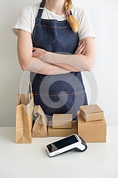 Young woman wearing apron with the arms crossed with carton boxes, cardboard packages and modern payment terminal at the