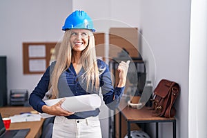 Young woman wearing architect hardhat pointing thumb up to the side smiling happy with open mouth