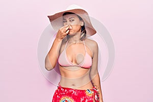 Young woman wearing bikini and hat smelling something stinky and disgusting, intolerable smell, holding breath with fingers on