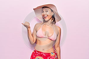 Young woman wearing bikini and hat smiling with happy face looking and pointing to the side with thumb up