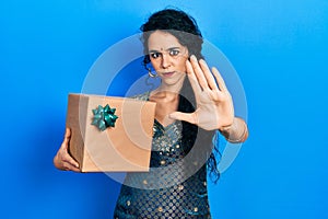 Young woman wearing bindi and traditional kurta dress holding gift with open hand doing stop sign with serious and confident