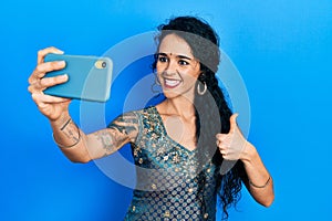 Young woman wearing bindi and traditional kurta dress taking a selfie photo with smartphone smiling happy and positive, thumb up