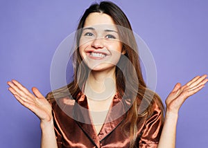 Young woman wearing brown pajama smiling, laughing, raising hands to up over purple background