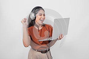 Young woman wearing brown shirt and headphone while holding laptop to listen music with clenched fist gesture isolated over white