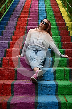 Young woman wearing casual clothes and sunglasses sits on rainbow painted stairs