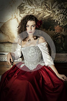 A young woman wearing a corset and red skirt and sitting in an opulent, vintage setting