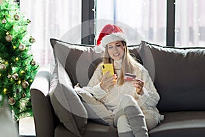 Young woman wearing cozy knitted sweater and red Santa hat when Paying online with credit card at home, holiday shopping