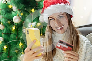 Young woman wearing cozy knitted sweater and red Santa hat when Paying online with credit card at home, holiday shopping