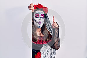 Young woman wearing day of the dead custome holding blank empty banner amazed and surprised looking up and pointing with fingers