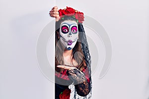 Young woman wearing day of the dead custome holding blank empty banner surprised pointing with finger to the side, open mouth