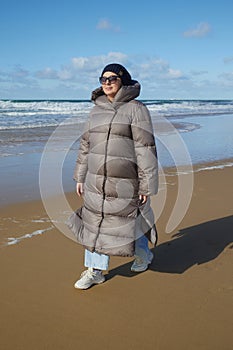 A young woman wearing a down jacket enjoys the warmth of the sun while walking along the sandy ocean beach