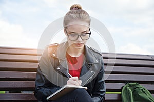 Young woman wearing eyeglasses writing notes on notebook with pen outside, planning day