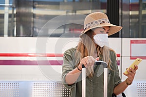 Young woman wearing face mask and explorer hat, using her smartphone while is waiting for the train at a train station
