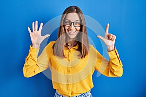 Young woman wearing glasses standing over blue background showing and pointing up with fingers number seven while smiling