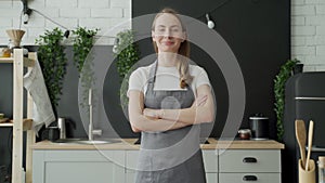 Young woman wearing gray apron smiling and crossing arms standing in her kitchen.