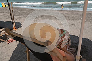 Young woman wearing hat and dress resting on a wooden chair on the beach with the sea in the background during a sunny morning