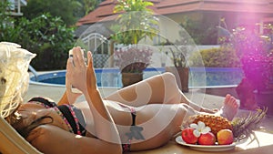 Young woman wearing hat lies on a sunbed uses smart phone next to the swimming pool on summer holidays with beautiful