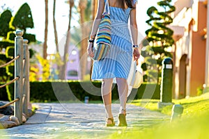 Young woman wearing light blue summer dress holding fashionable shoulder bag and yellow straw hat standing outside enjoying warm