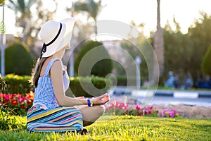 Young woman wearing light blue summer dress and yellow straw hat relaxing on green grass lawn in summer park. Girl in casual