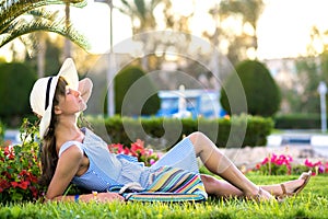 Young woman wearing light blue summer dress and yellow straw hat relaxing on green grass lawn in summer park. Girl in casual