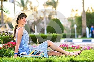 Young woman wearing light blue summer dress and yellow straw hat relaxing on green grass lawn in summer park. Girl in casual