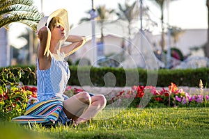 Young woman wearing light blue summer dress and yellow straw hat relaxing on green grass lawn in summer park. Girl in casual