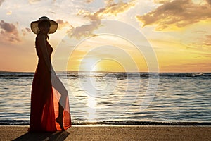 Young woman wearing long red dress and straw hat standing on sand beach at sea shore enjoying view of rising sun in early summer