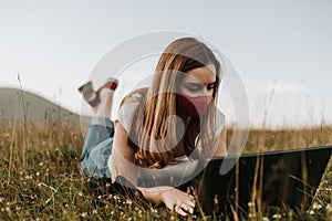 Young women with mask lies in the grass and looking at laptop
