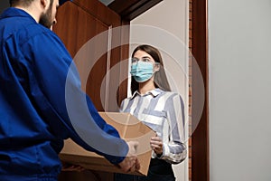 Young woman wearing mask receiving parcel from delivery man indoors. Prevention of virus spread